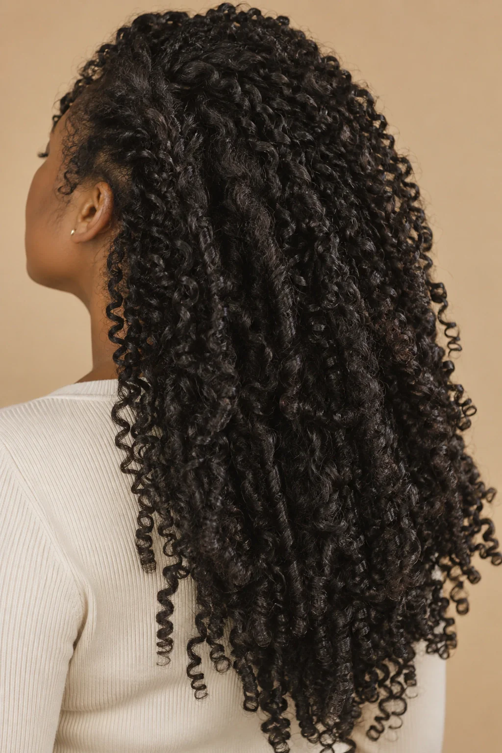 A close side and back view of a young Black woman’s long, defined natural coils against a warm neutral background.